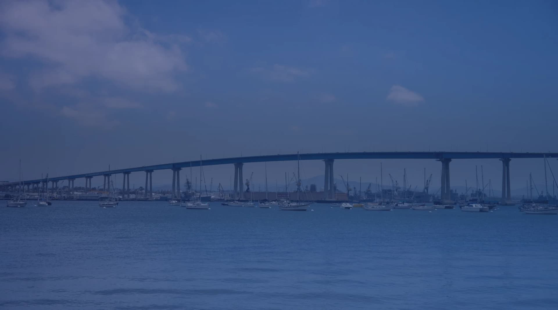 A long curved bridge stretching across a calm bay, with sailboats and ships moored along the far shore