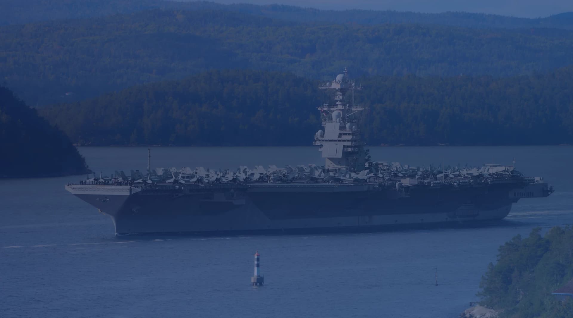 A Navy aircraft carrier with jets parked on its flight deck moving through a calm channel lined with wooded hills