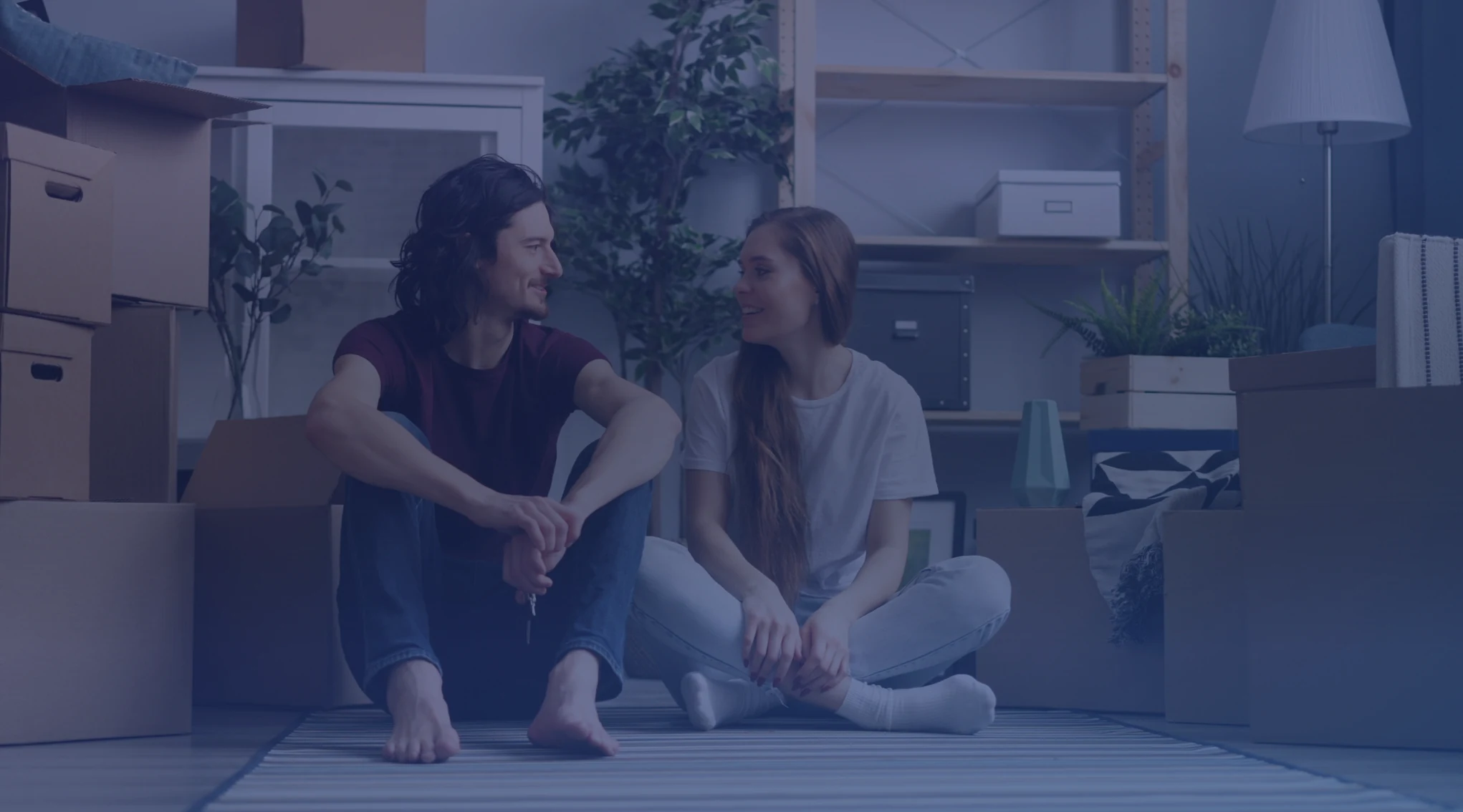 A couple sitting on the floor of a new home surrounded by moving boxes while they wait