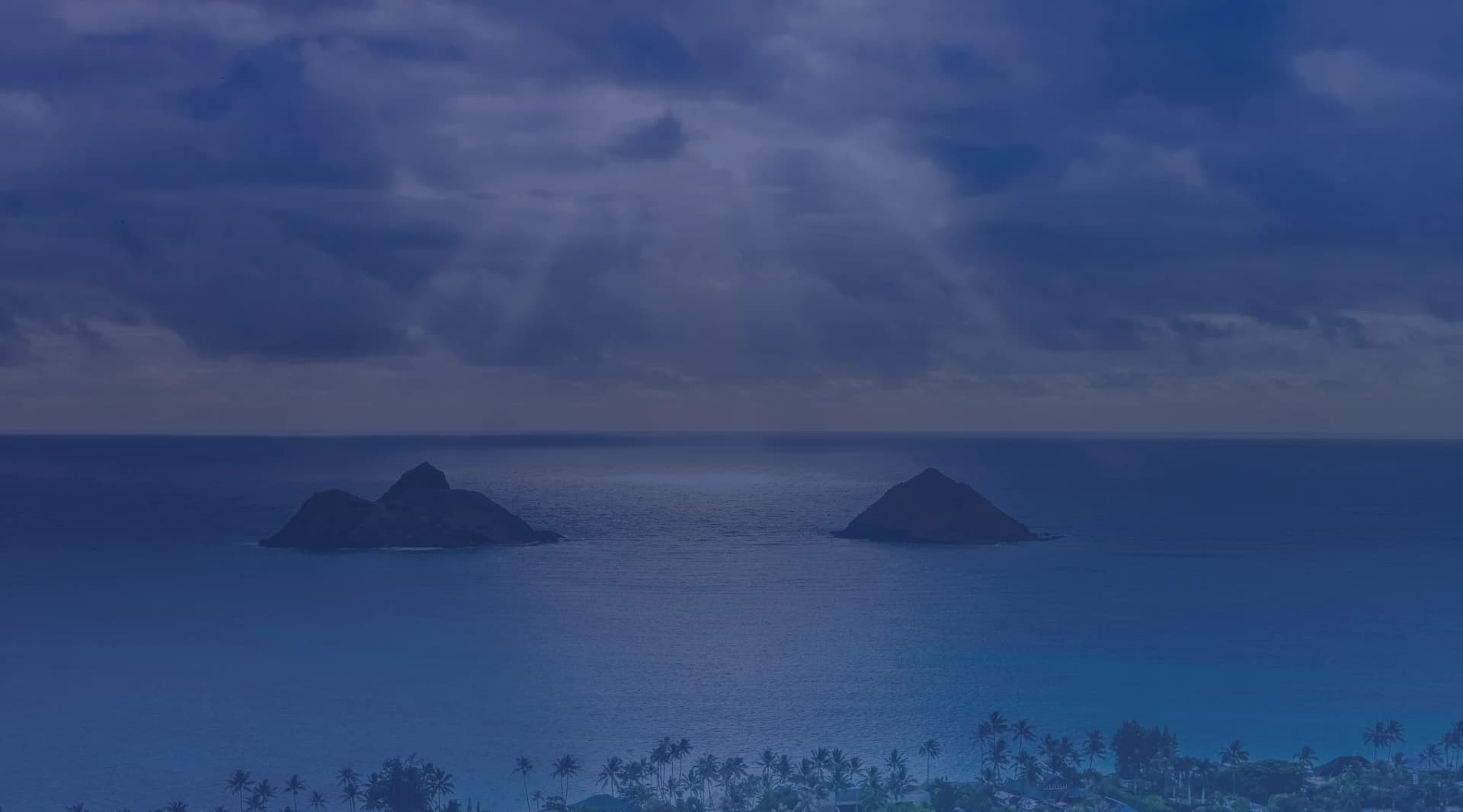 The Mokulua Islands rising from turquoise water off Lanikai Beach on windward Oahu, with sun rays breaking through dramatic clouds above the Pacific