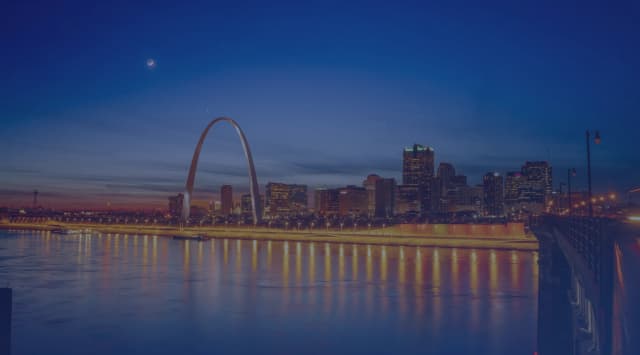 St. Louis skyline at dusk viewed from across the Mississippi River, with the Gateway Arch rising above the city and the Illinois Metro East shore in the foreground
