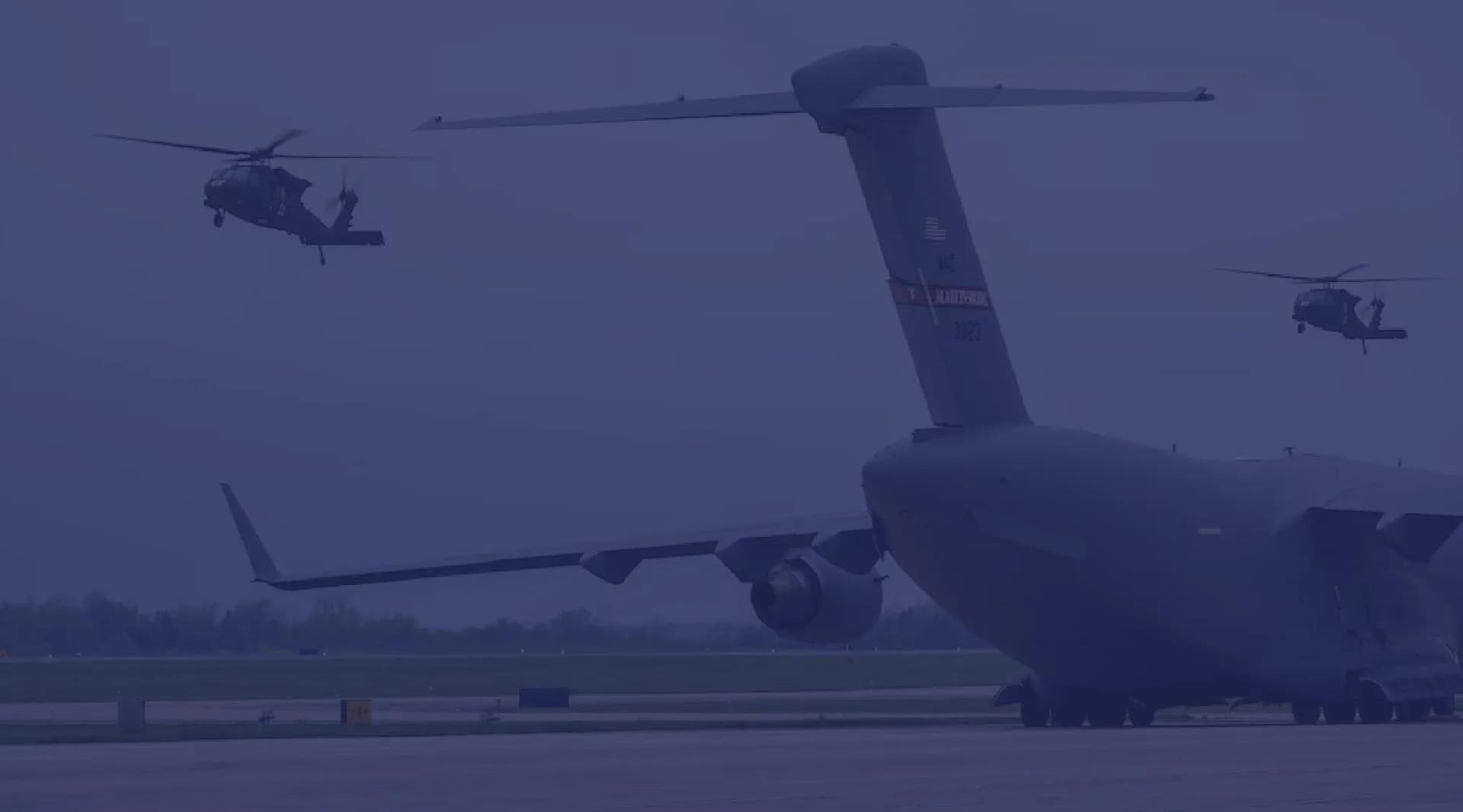 A C-5 Galaxy cargo plane sits on the runway in West Virginia while AH-64 Apaches fly around it.