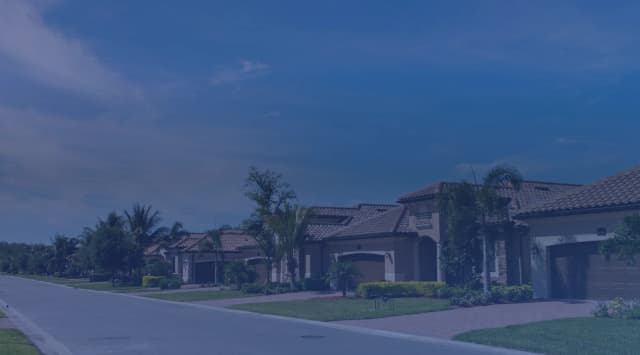A quiet suburban street with palm trees, tile-roofed houses, and green lawns under a wide sky.