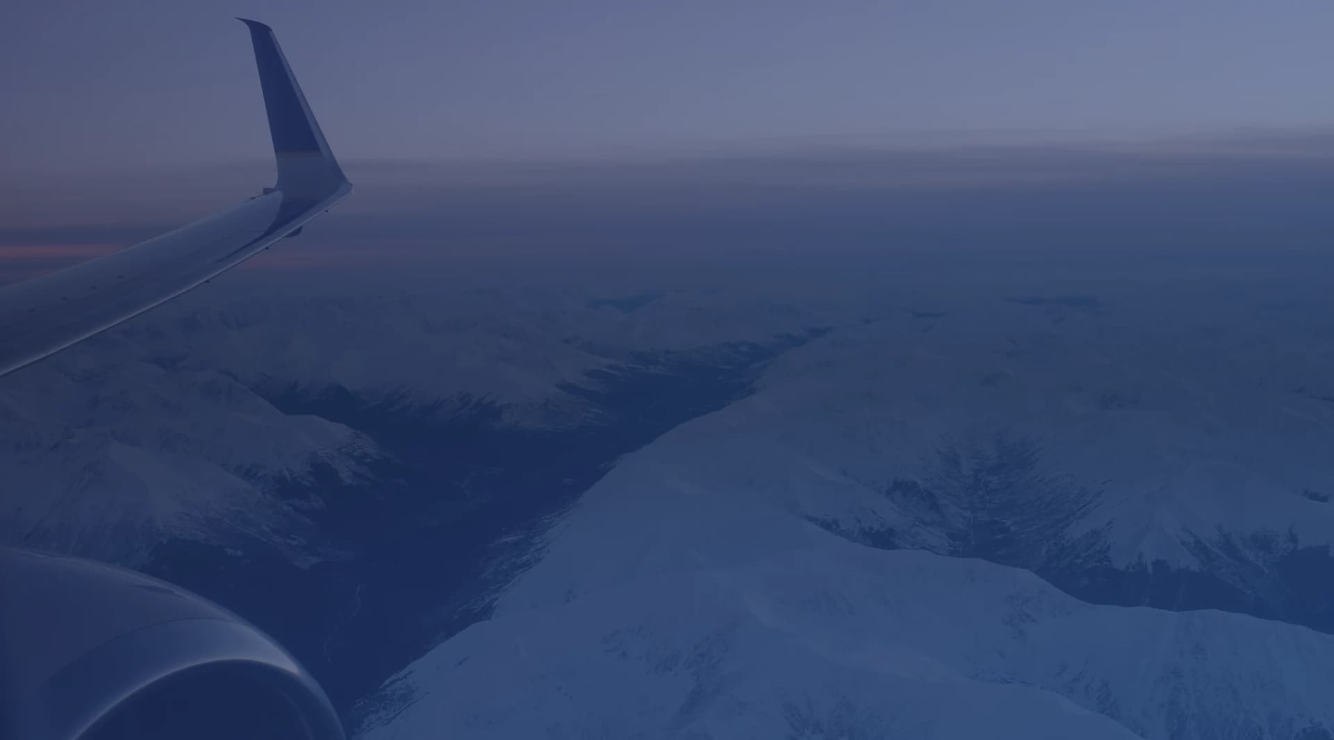 Snow-covered mountain ranges seen from an aircraft window, with the wing in the foreground