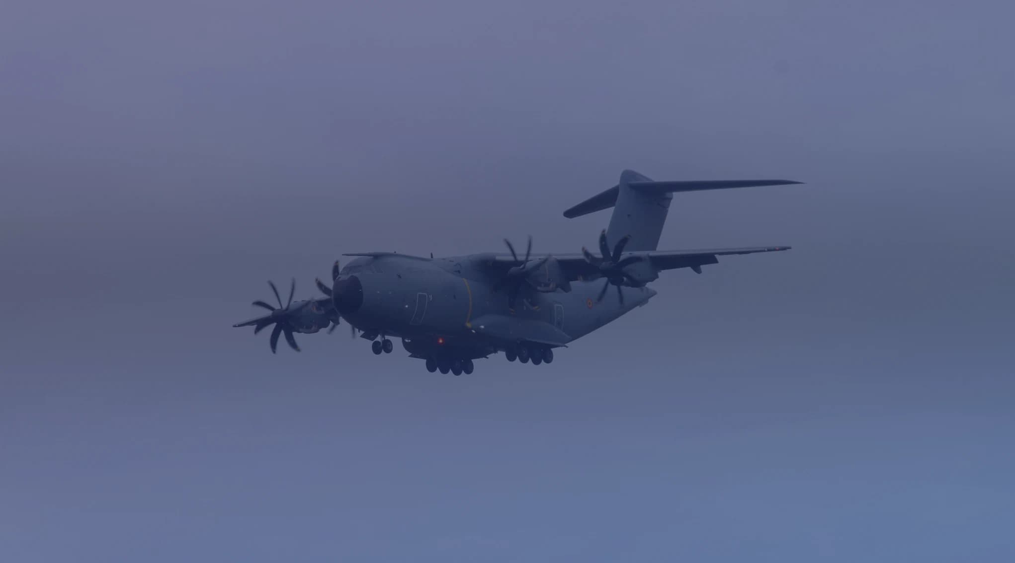 A four-engine military transport aircraft flying low through a hazy overcast sky