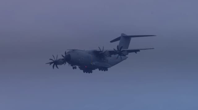 A four-engine military transport aircraft flying low through a hazy overcast sky