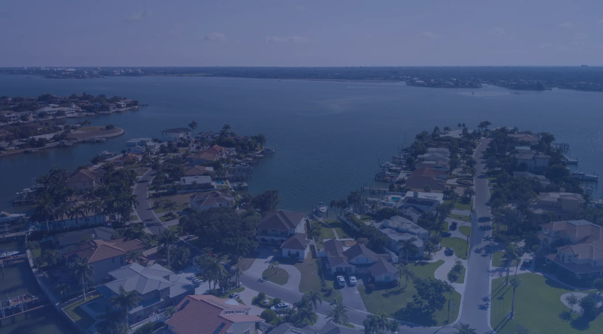 A quiet residential street of single-story homes with palm trees and a Gulf Coast inlet visible in the distance