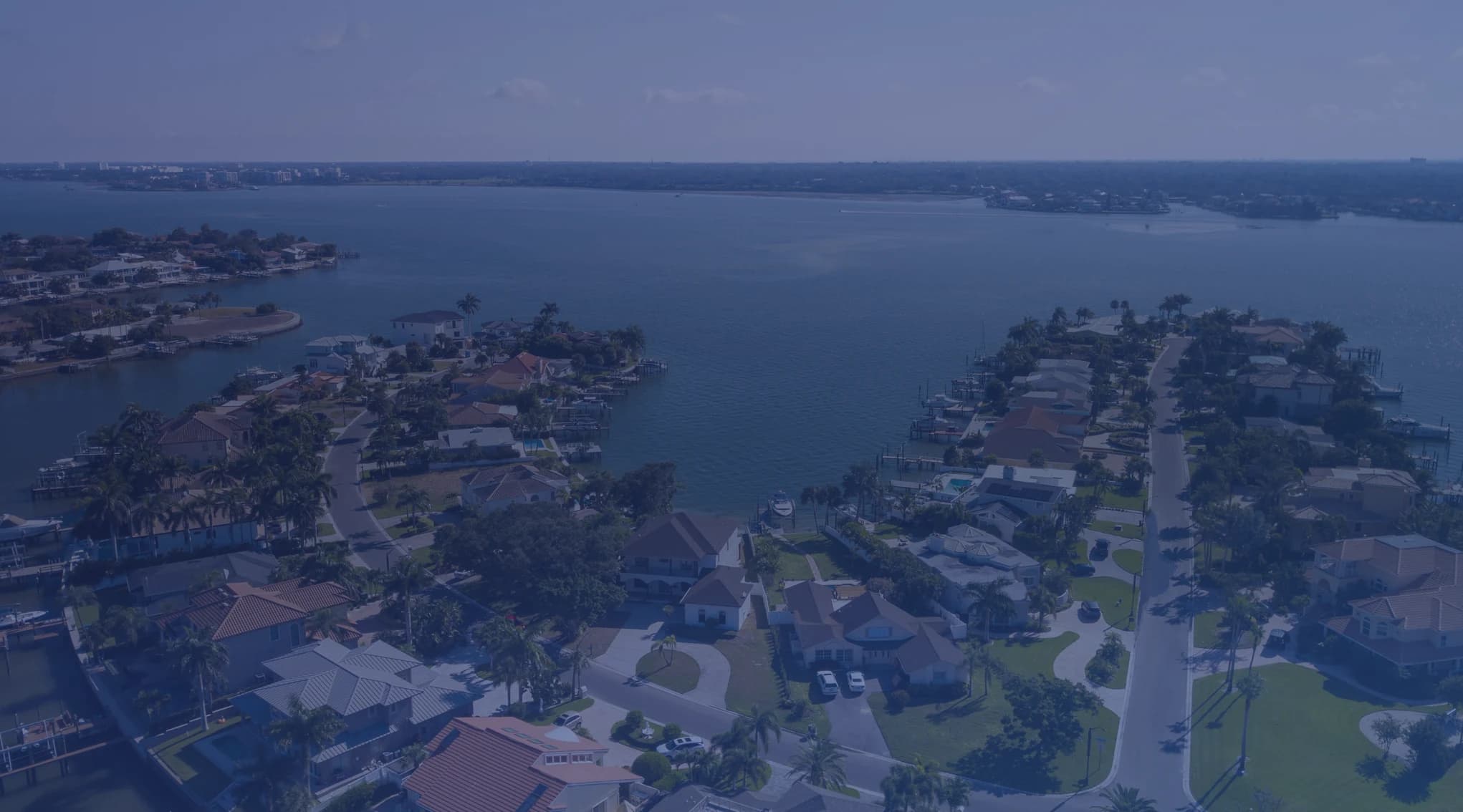 A quiet residential street of single-story homes with palm trees and a Gulf Coast inlet visible in the distance