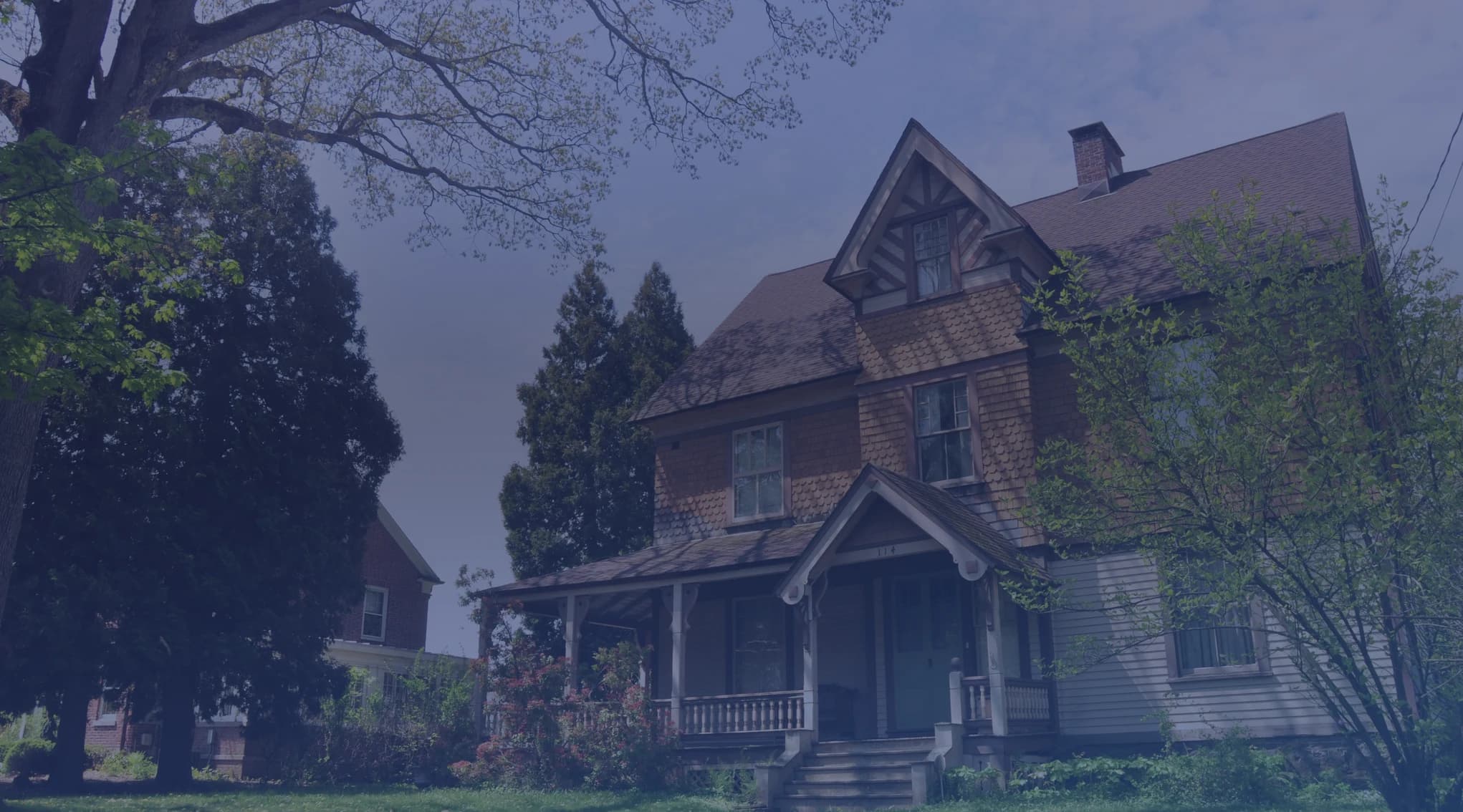 A two-story shingled home with a covered front porch, framed by mature shade trees on a quiet residential street