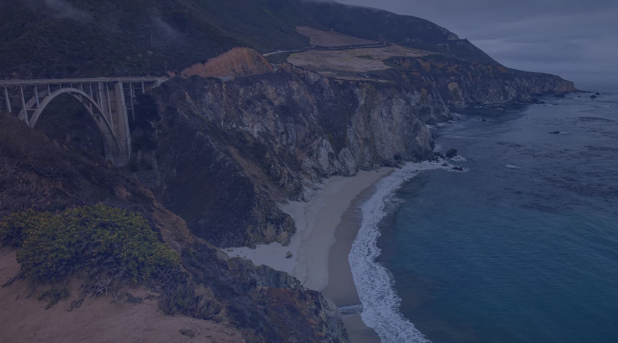 The rugged Big Sur coastline in California, with a cove, cliffs, and the Pacific Ocean under a cloudy sky.