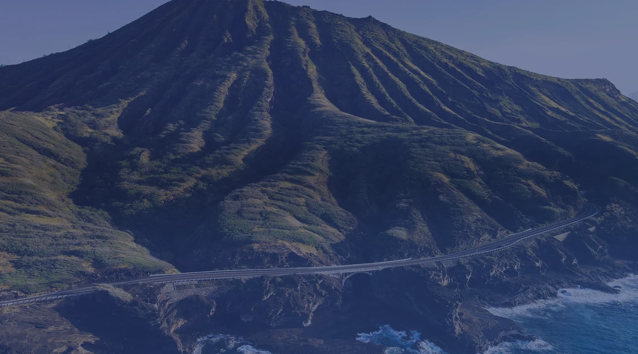 An aerial view of a green coastal mountain on Oahu, Hawaii, with a road curving along the shoreline above the ocean.