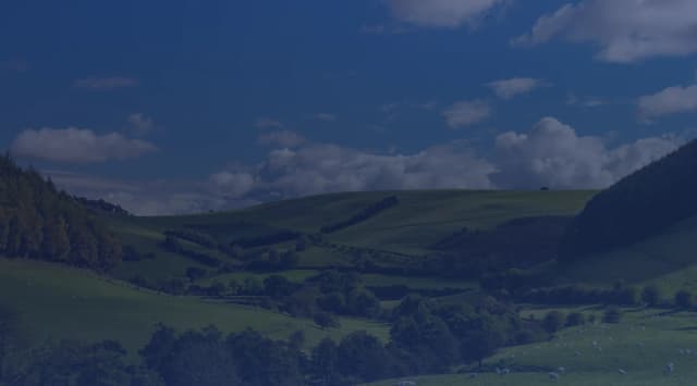 Lush green rolling hills and a hedge-lined valley under a blue sky dotted with puffy white clouds.