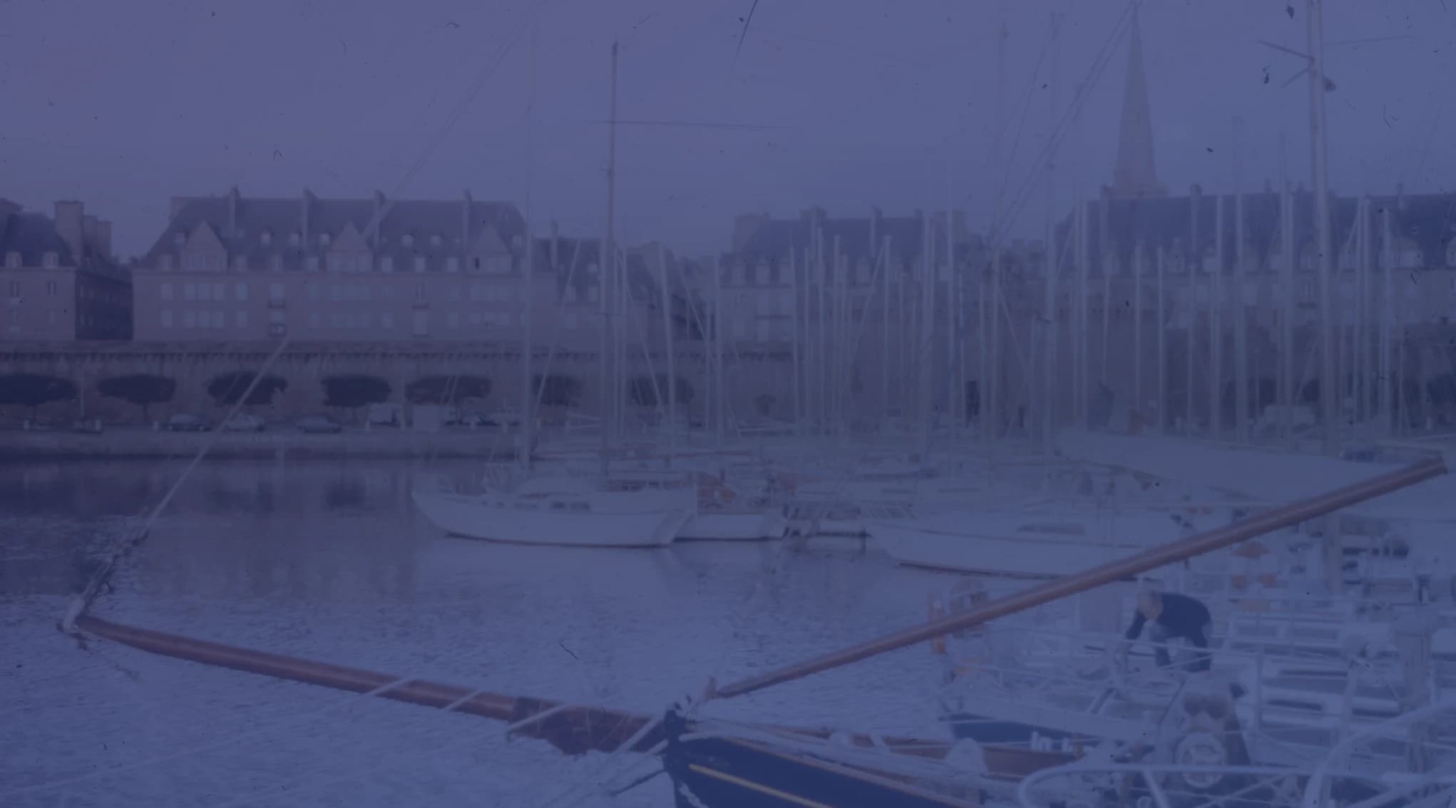 Sailboats moored in a calm waterfront harbor below stone buildings and a church steeple.