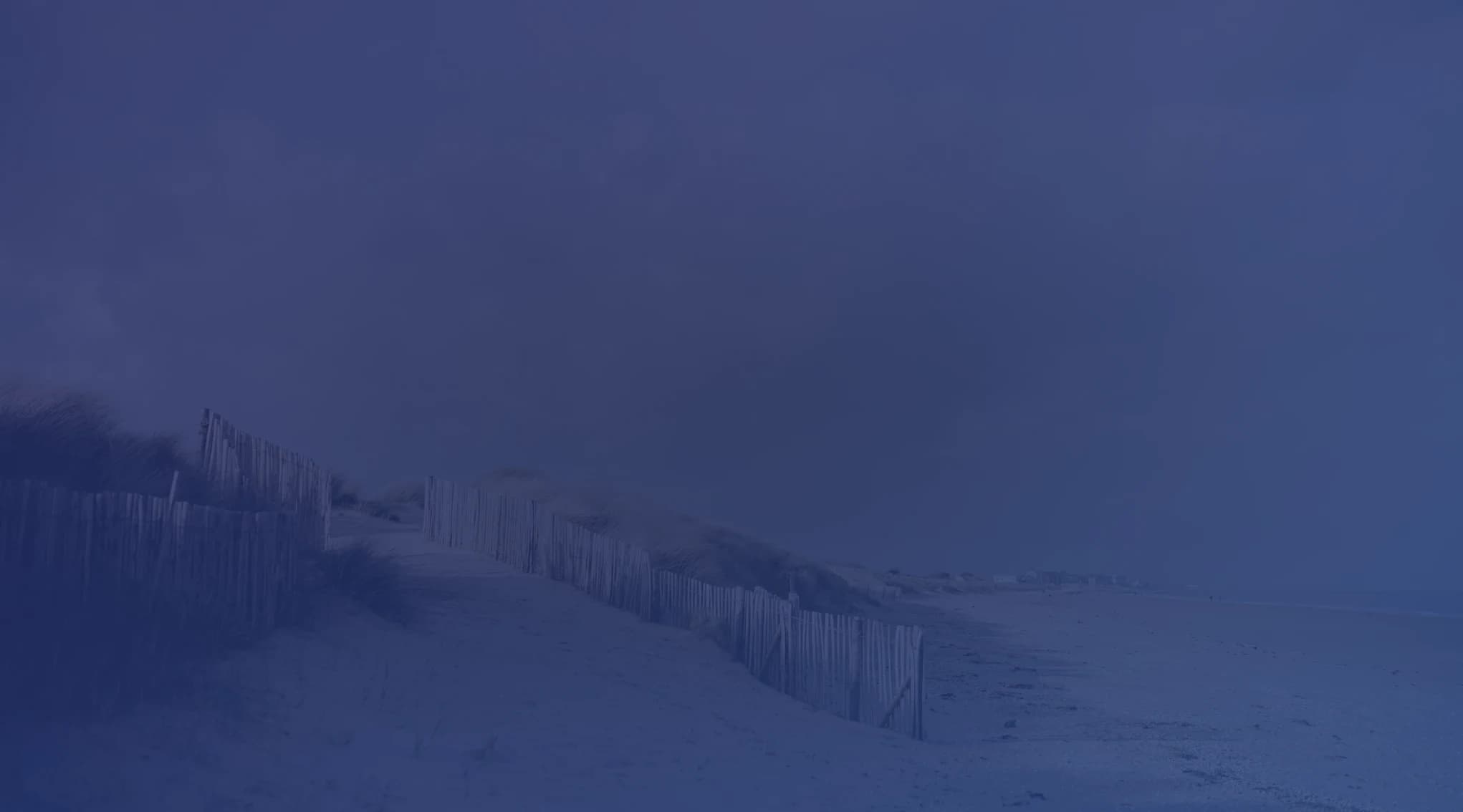 A weathered wooden fence runs over low grassy sand dunes toward a wide, empty beach under a soft, partly cloudy sky.