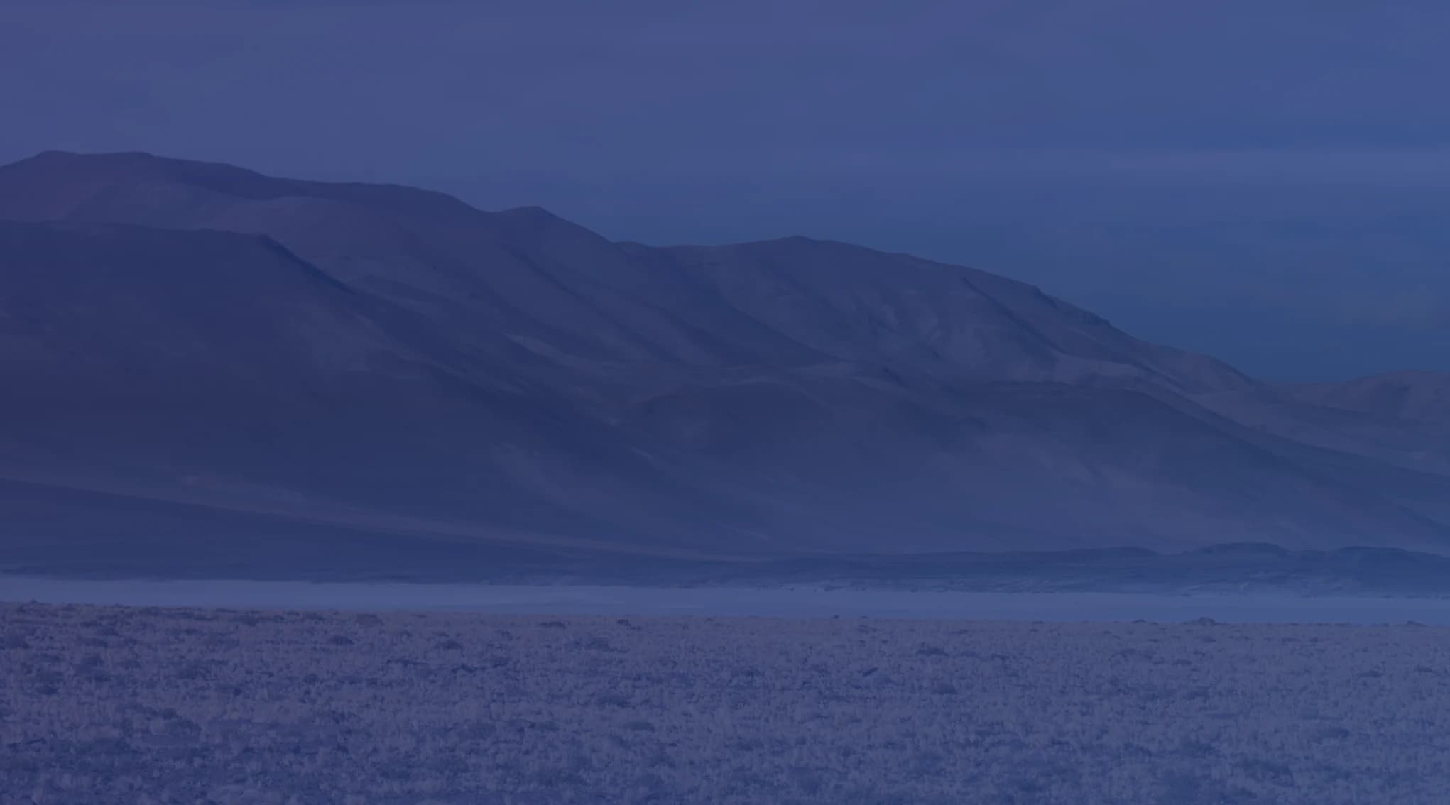 A dry, high plateau of golden grass tufts and reddish soil below a barren mountain ridge under a blue sky.