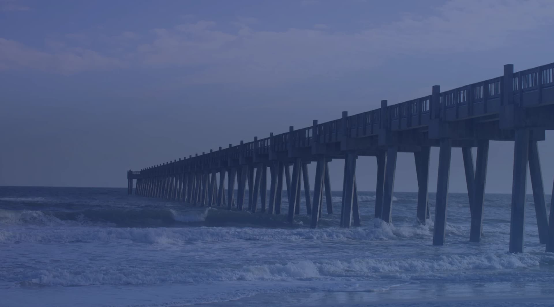 A long wooden fishing pier stretching out over rolling surf under a cloudy sky on a Florida Gulf Coast beach