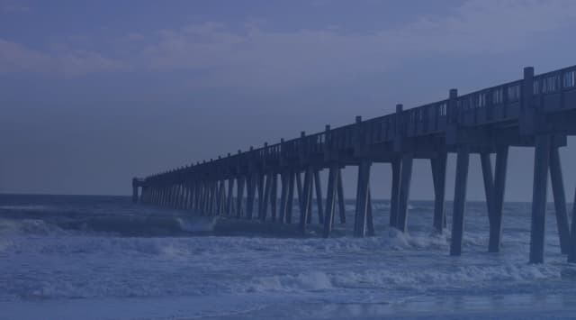 A long wooden fishing pier stretching out over rolling surf under a cloudy sky on a Florida Gulf Coast beach