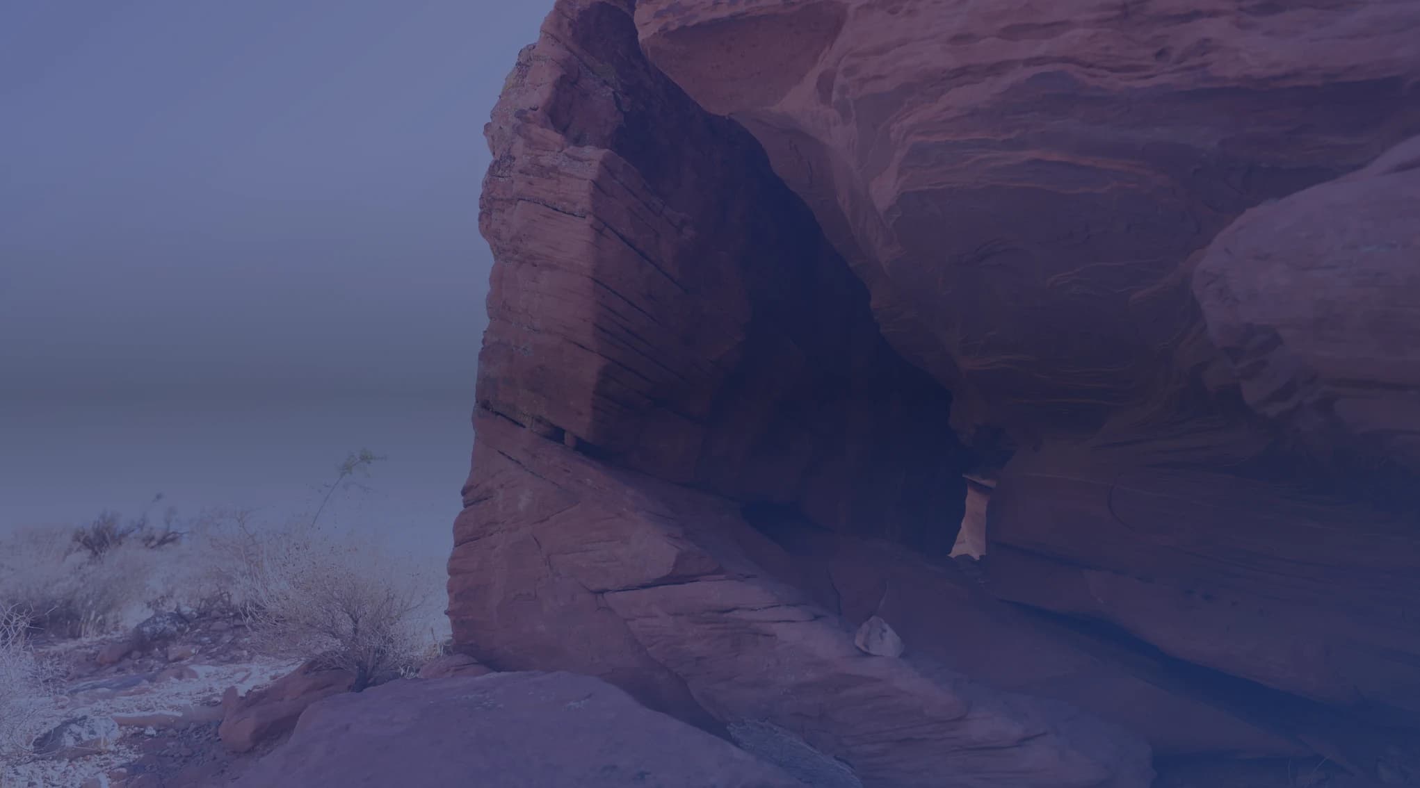 A large red sandstone rock face and overhang rise above desert scrub under a clear blue sky.