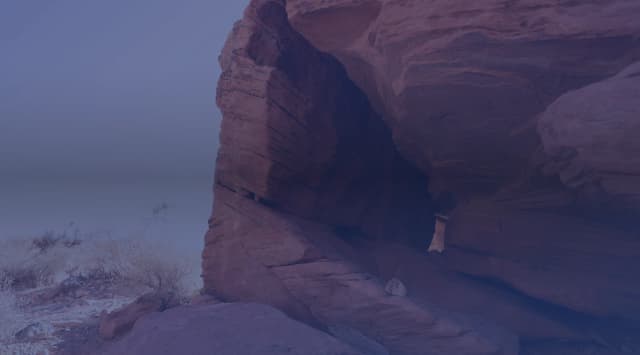 A large red sandstone rock face and overhang rise above desert scrub under a clear blue sky.