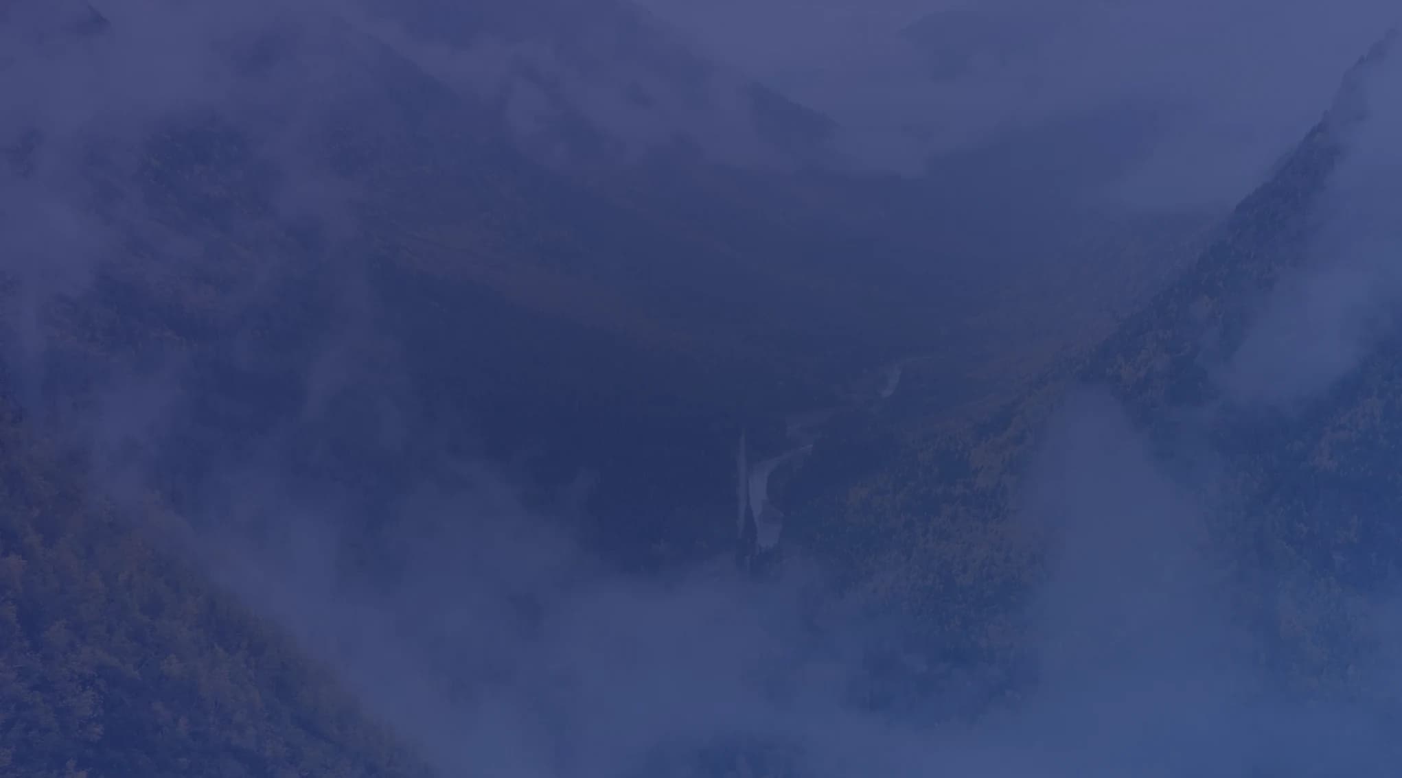Low clouds drift through a forested mountain valley touched with autumn yellow, with a river winding along the valley floor.