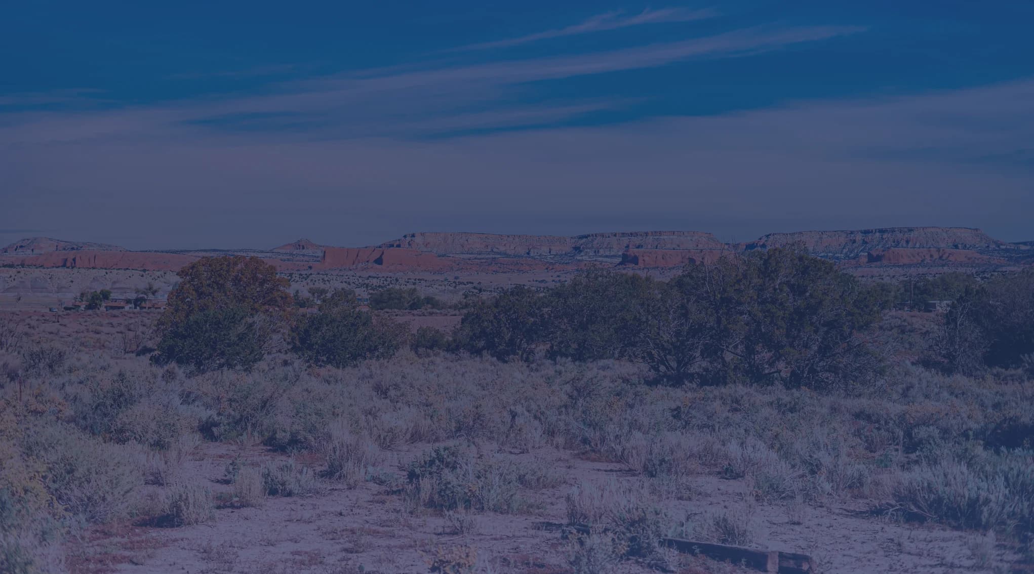 High-desert scrubland dotted with sagebrush stretches toward flat-topped red mesas under a wide blue sky.