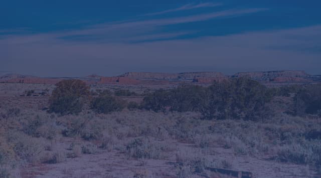 High-desert scrubland dotted with sagebrush stretches toward flat-topped red mesas under a wide blue sky.