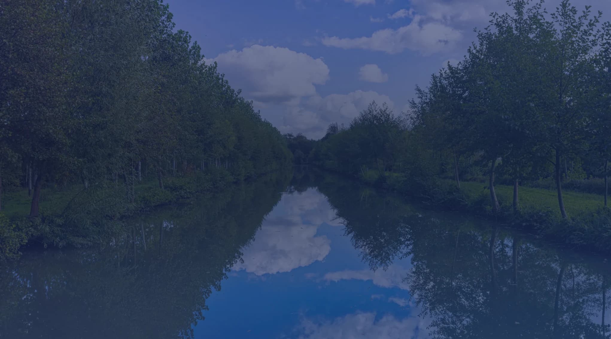 A calm tree-lined river reflects a blue, partly cloudy sky between green wooded banks.