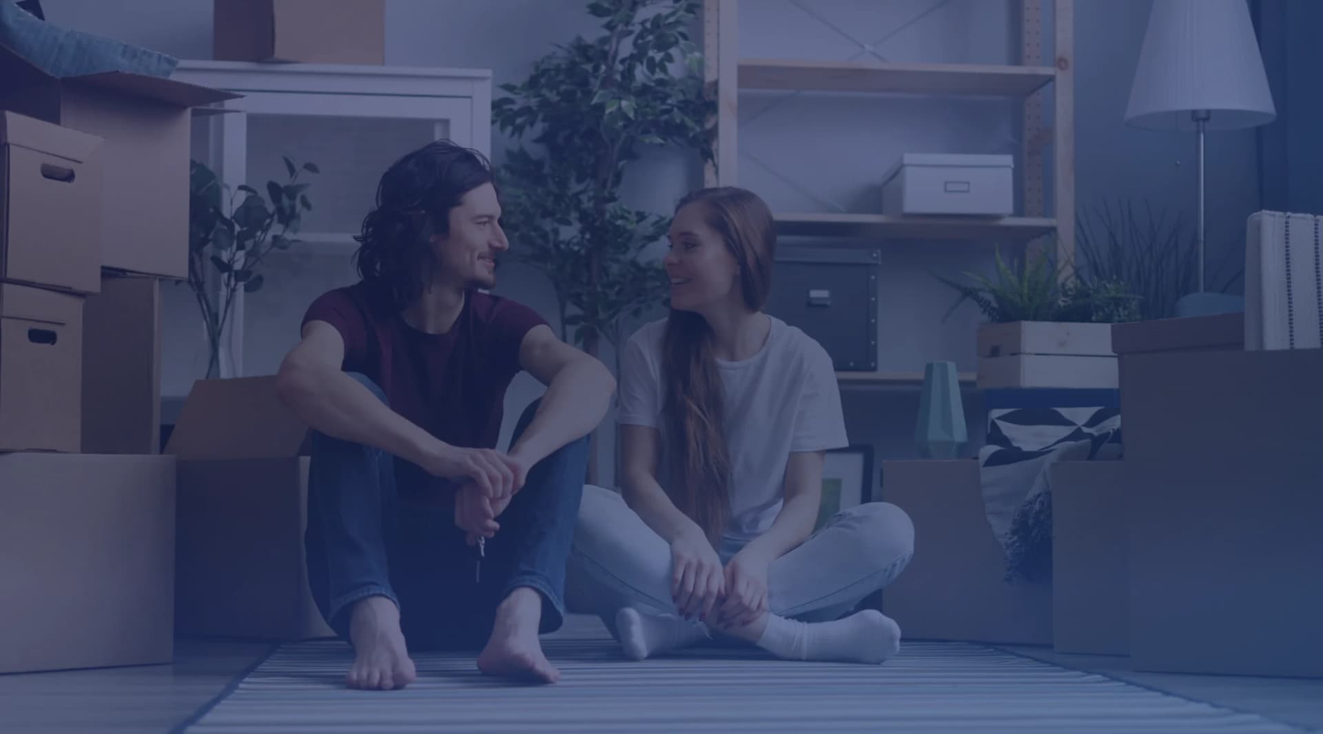 A couple sitting on the floor of a new home surrounded by moving boxes while they wait