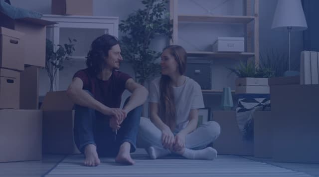 A couple sitting on the floor of a new home surrounded by moving boxes while they wait