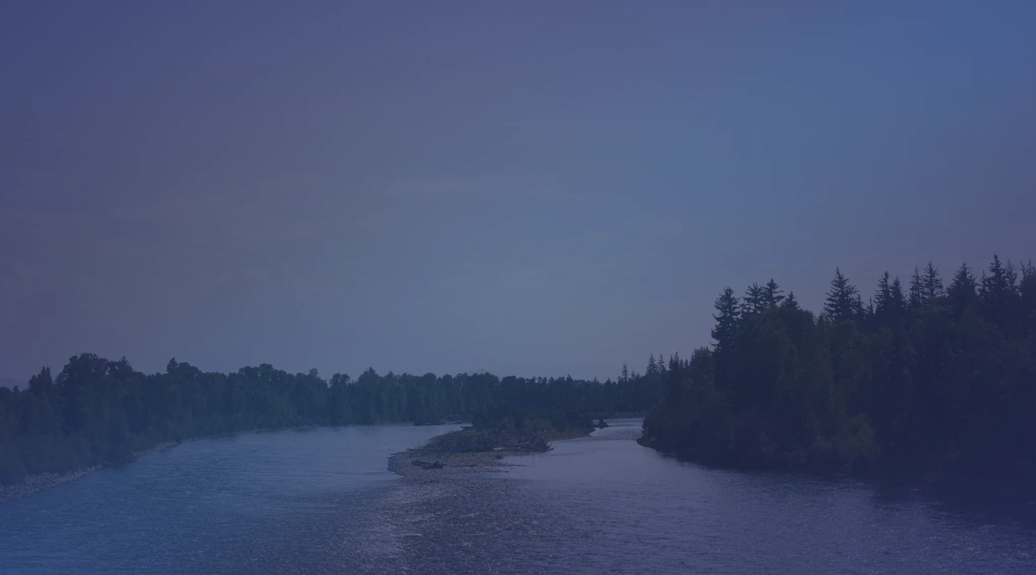 A calm, wide river bordered by green forested banks under a pale, hazy sky, like the wooded waterways of Northern Virginia.