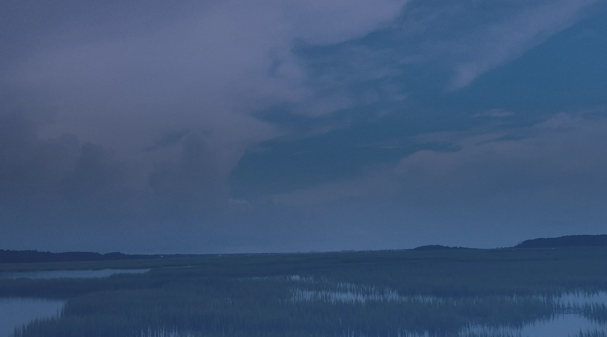A coastal salt marsh of tidal water and grass flats under a wide cloudy sky, like the Georgia Lowcountry near Fort Stewart and Savannah.