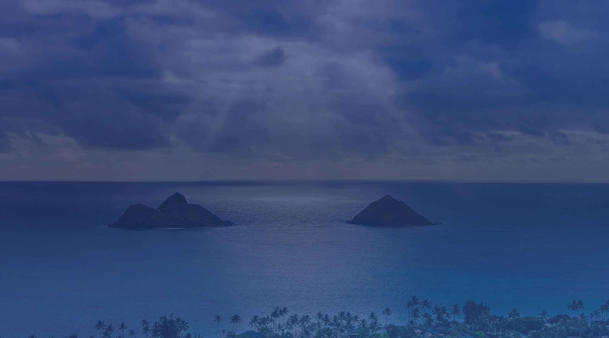 The Mokulua Islands rising from turquoise water off Lanikai Beach on windward Oahu, with sun rays breaking through dramatic clouds above the Pacific