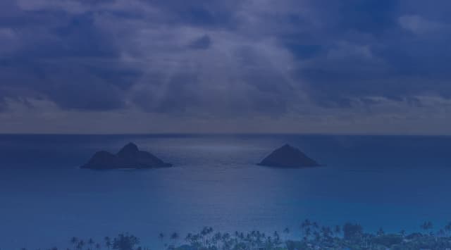 The Mokulua Islands rising from turquoise water off Lanikai Beach on windward Oahu, with sun rays breaking through dramatic clouds above the Pacific