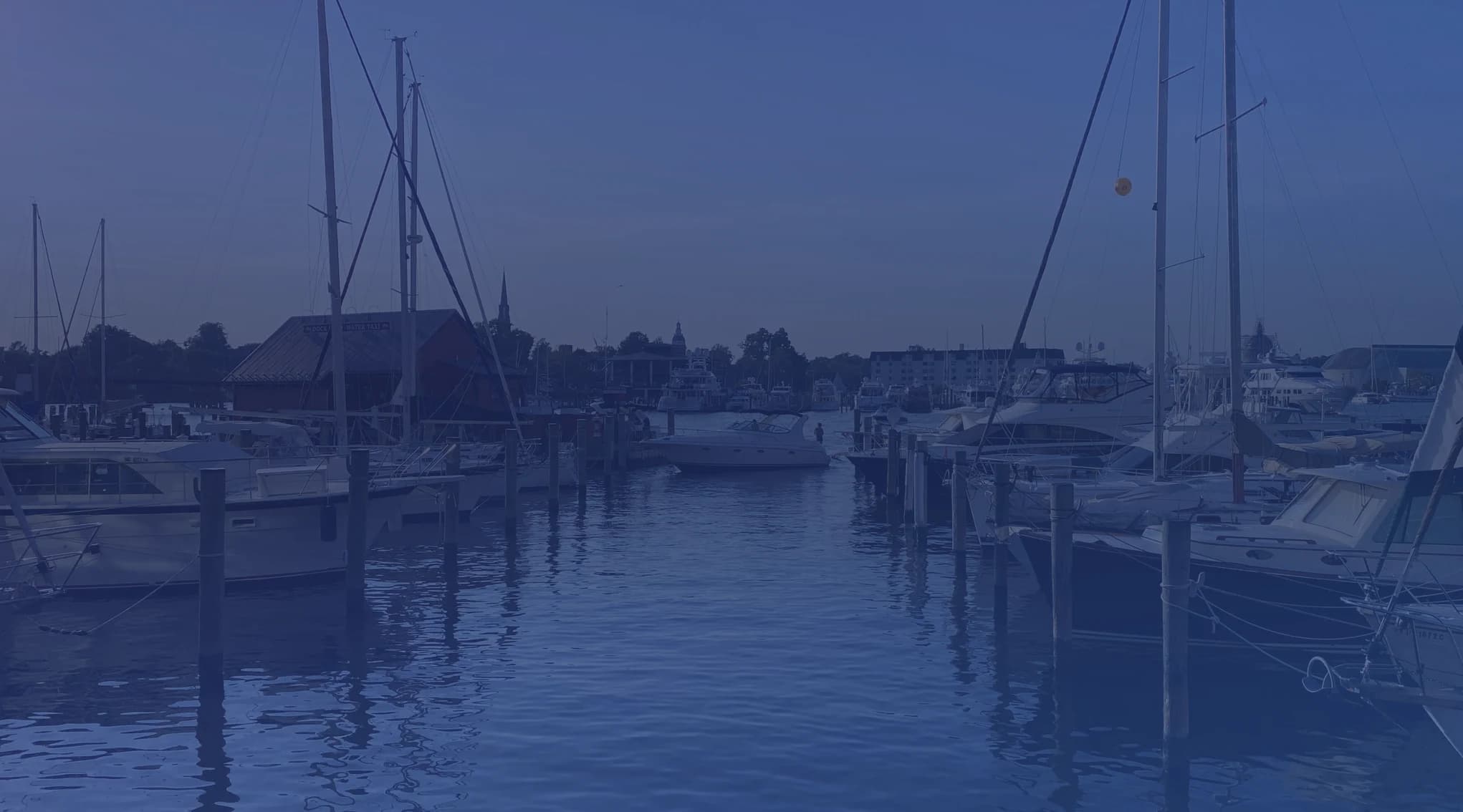 Sailboats and motorboats moored at an Annapolis marina with a church steeple and historic waterfront buildings visible across calm water under a clear blue sky