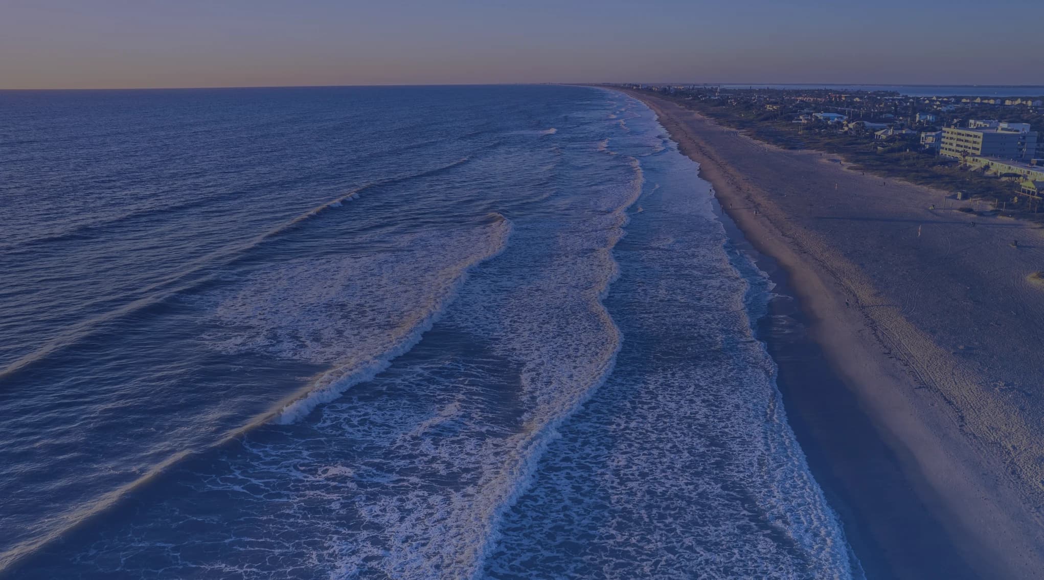 Aerial view of Cocoa Beach at sunrise, with Atlantic surf rolling onto a wide sandy shore and low-rise buildings lining the Florida Space Coast