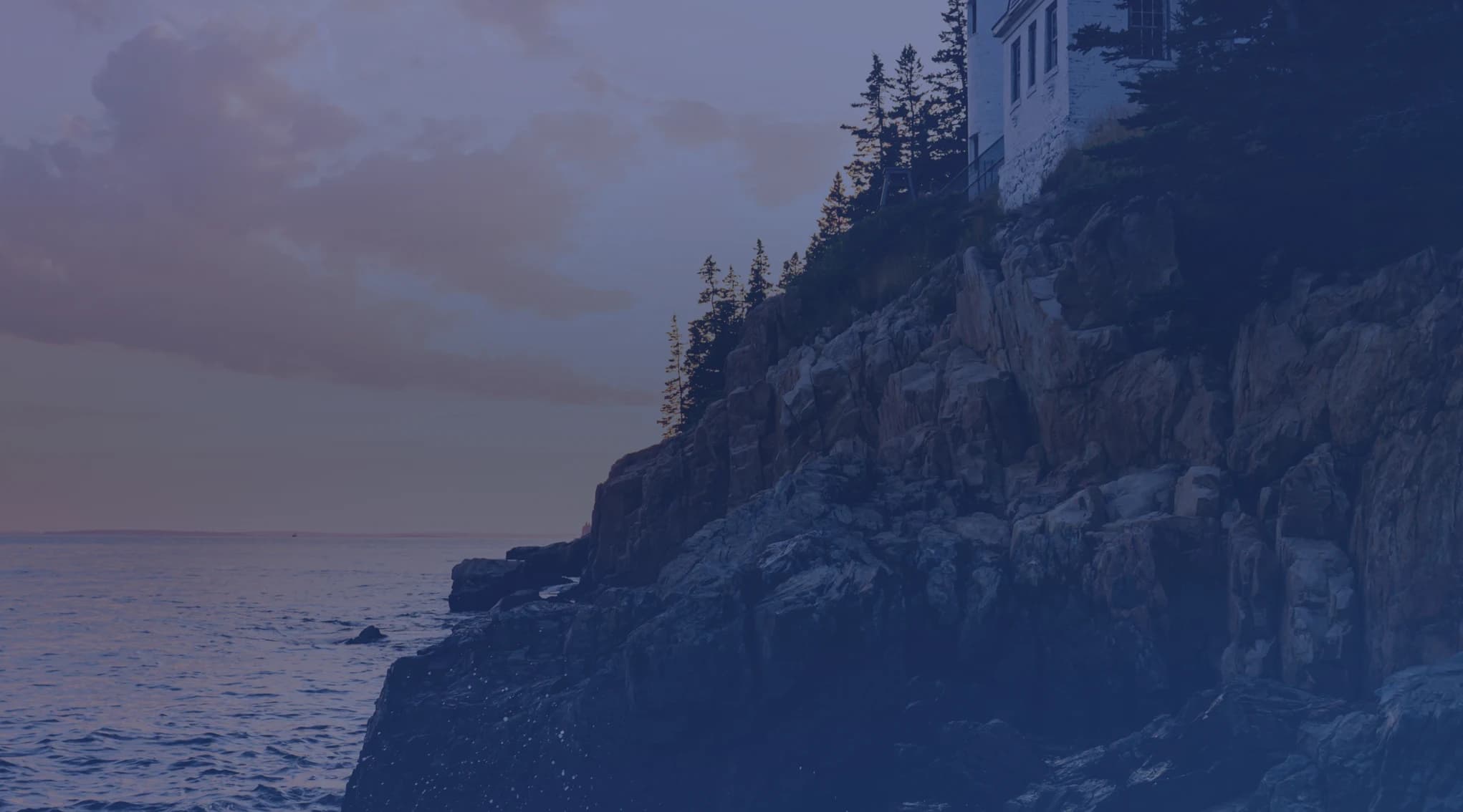 A lighthouse perched on rocky Maine coast cliffs at sunset, with the cold Atlantic Ocean stretching to the horizon