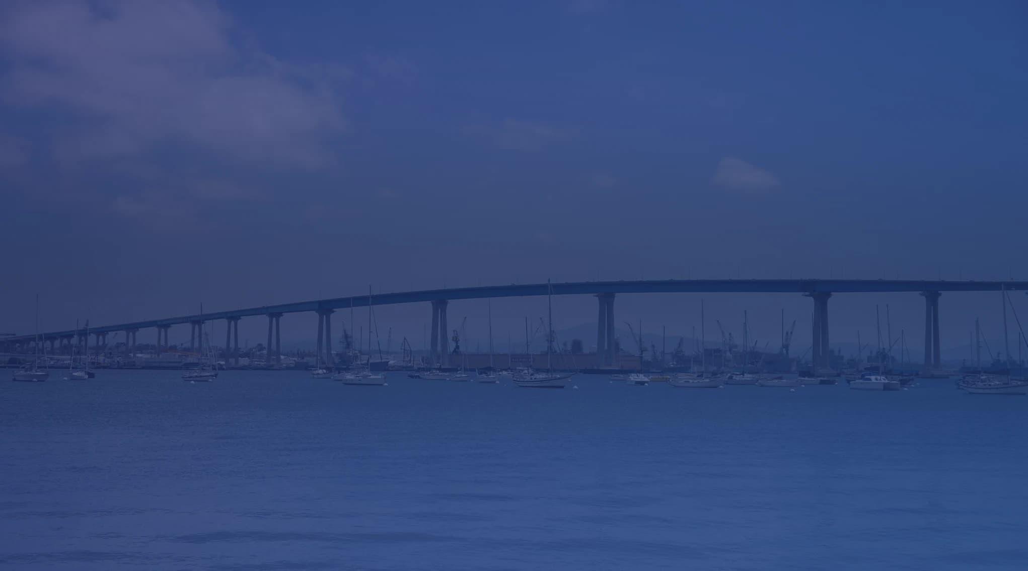 The Coronado Bridge spanning San Diego Bay with sailboats in the foreground and a bright blue sky above