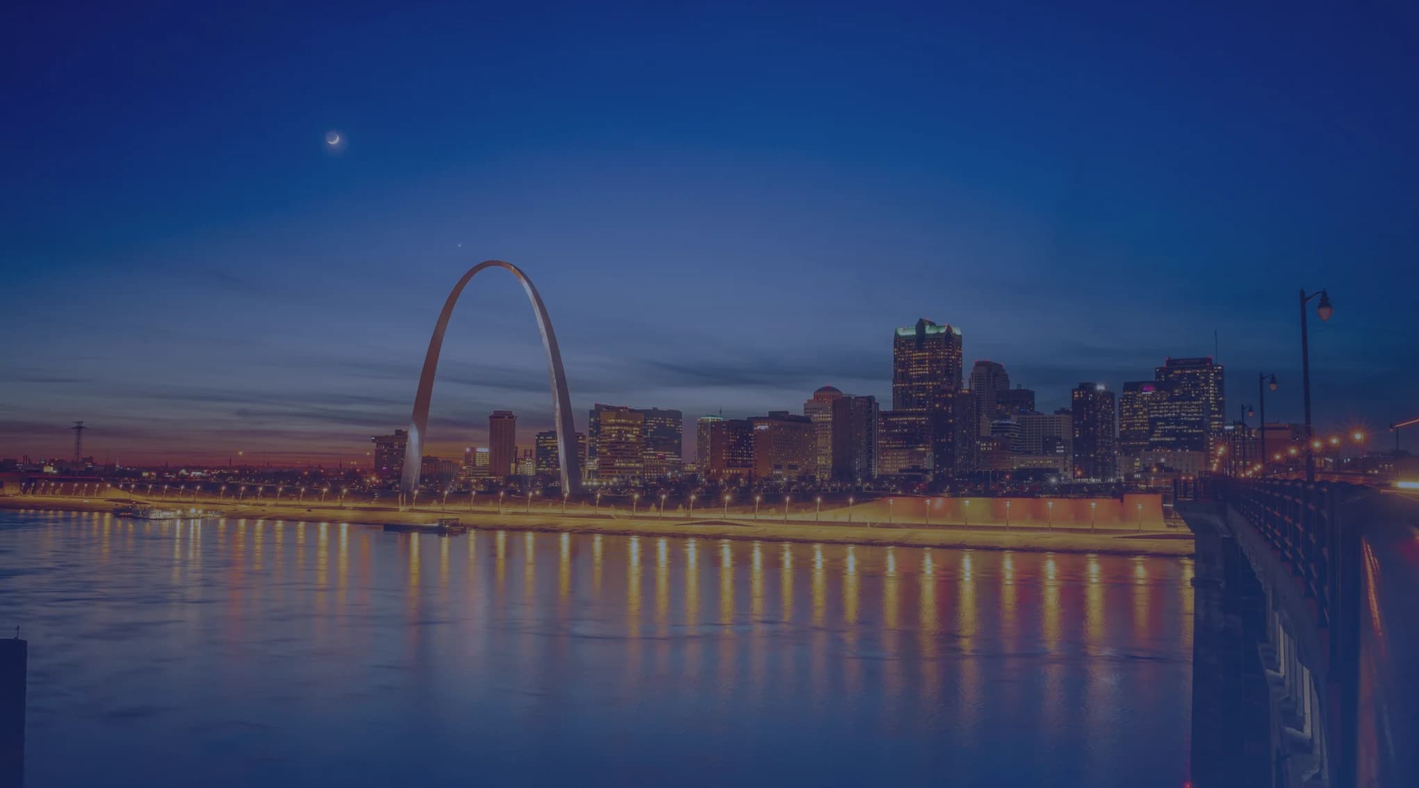 St. Louis skyline at dusk viewed from across the Mississippi River, with the Gateway Arch rising above the city and the Illinois Metro East shore in the foreground