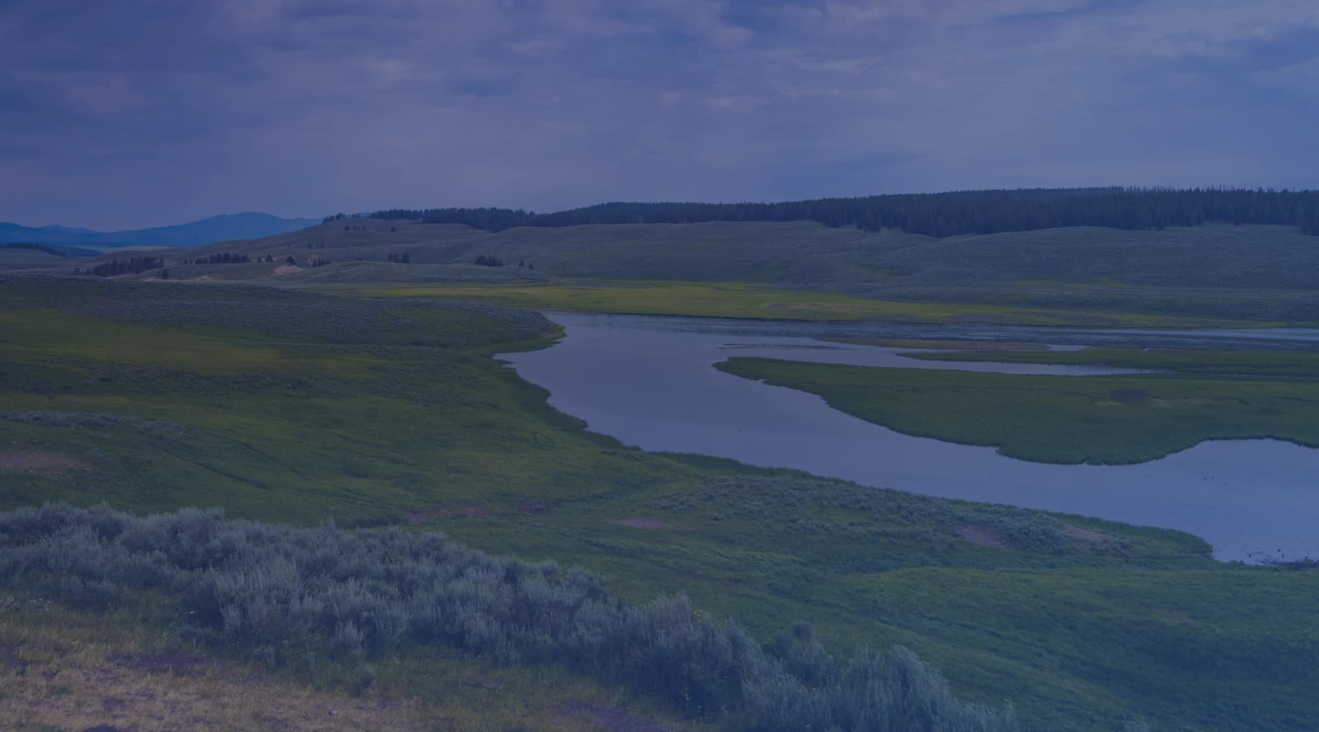 A river winding through a broad green valley under a cloudy summer sky.