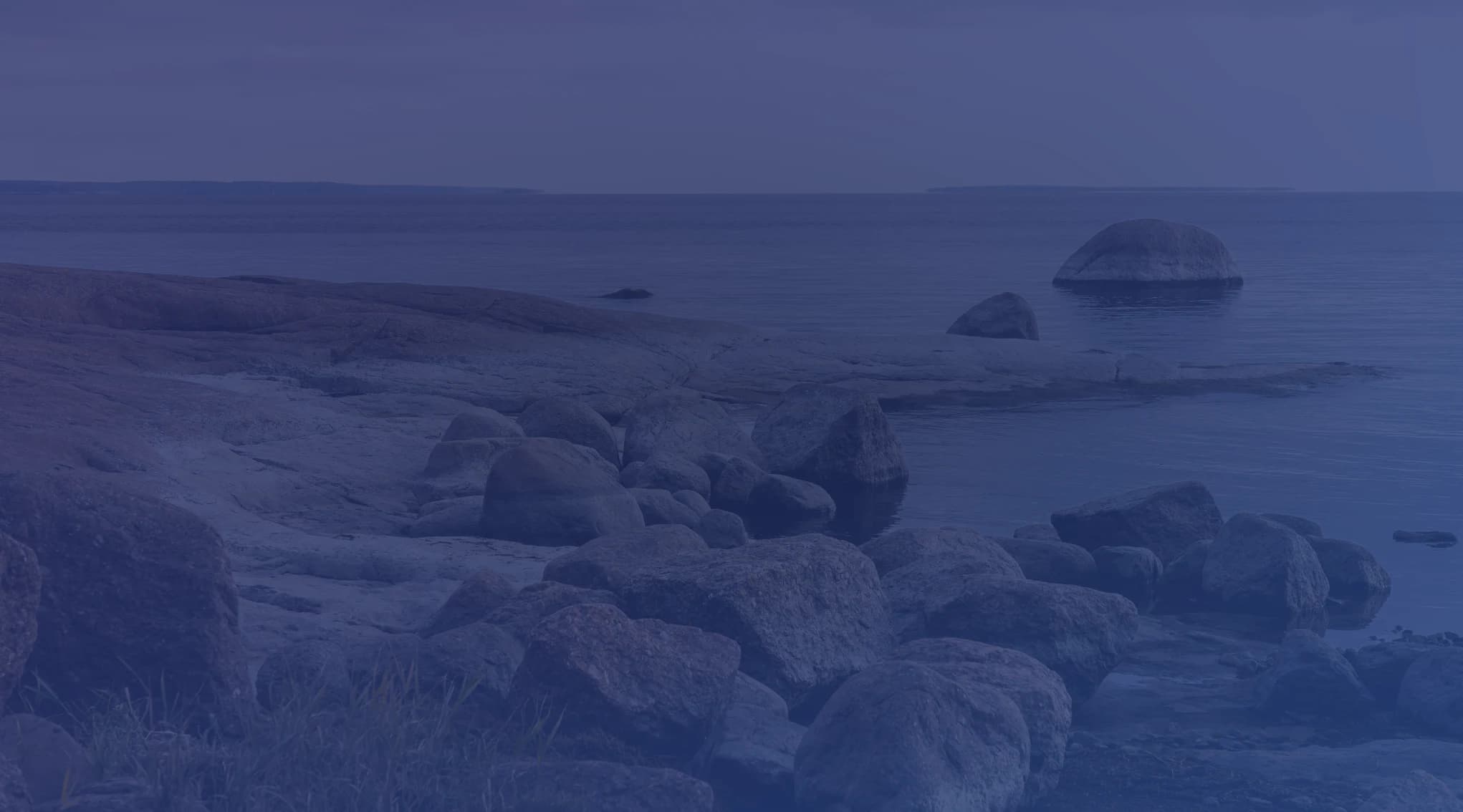 Large rounded boulders and a smooth rock shelf line a calm shoreline meeting flat gray water under an overcast sky.