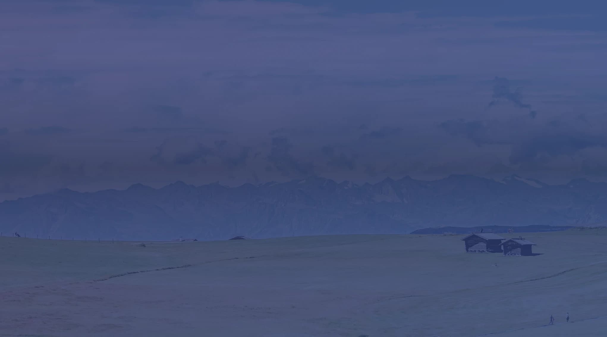 Rolling green pastureland with a small barn and a wooden fence, distant mountains on the horizon.