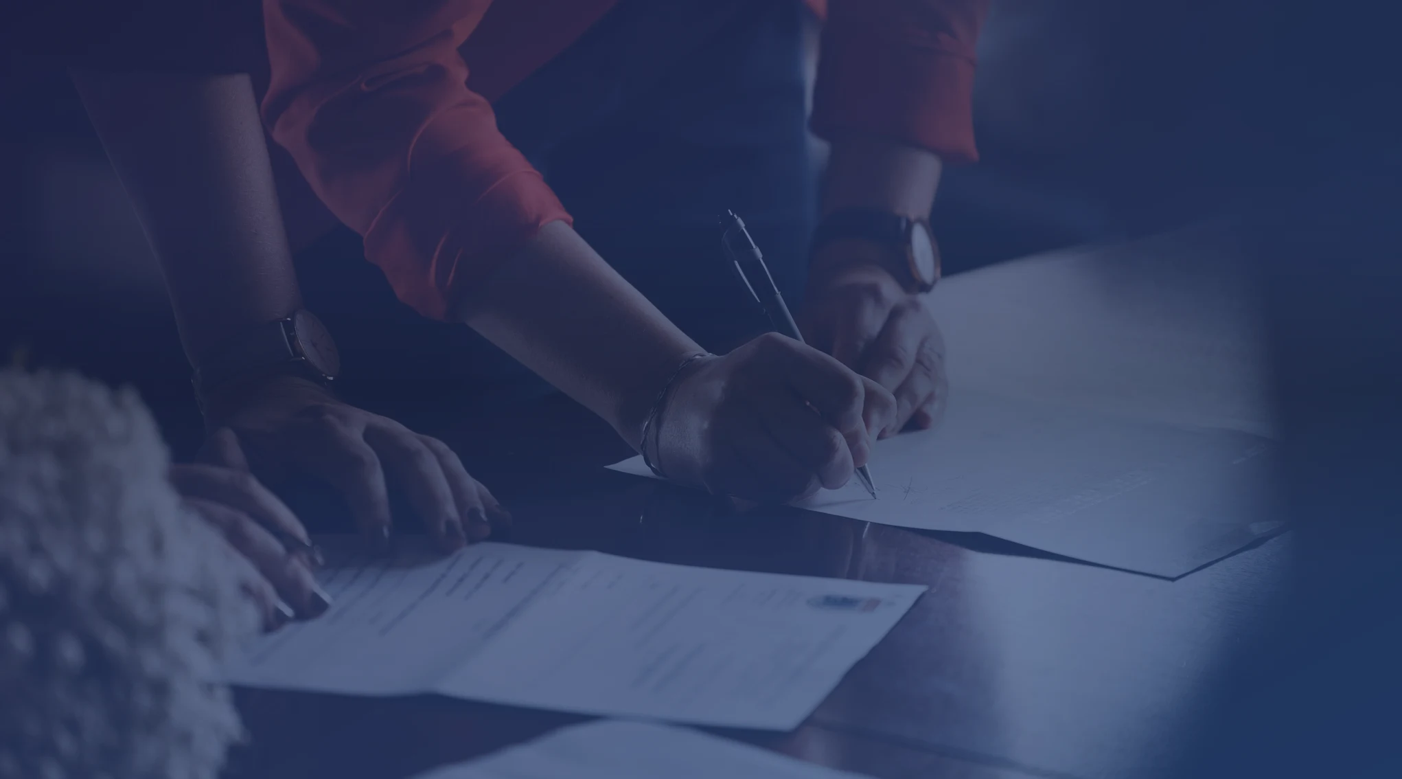 Two people at a table, one signing loan paperwork with a pen while the other holds the pages steady