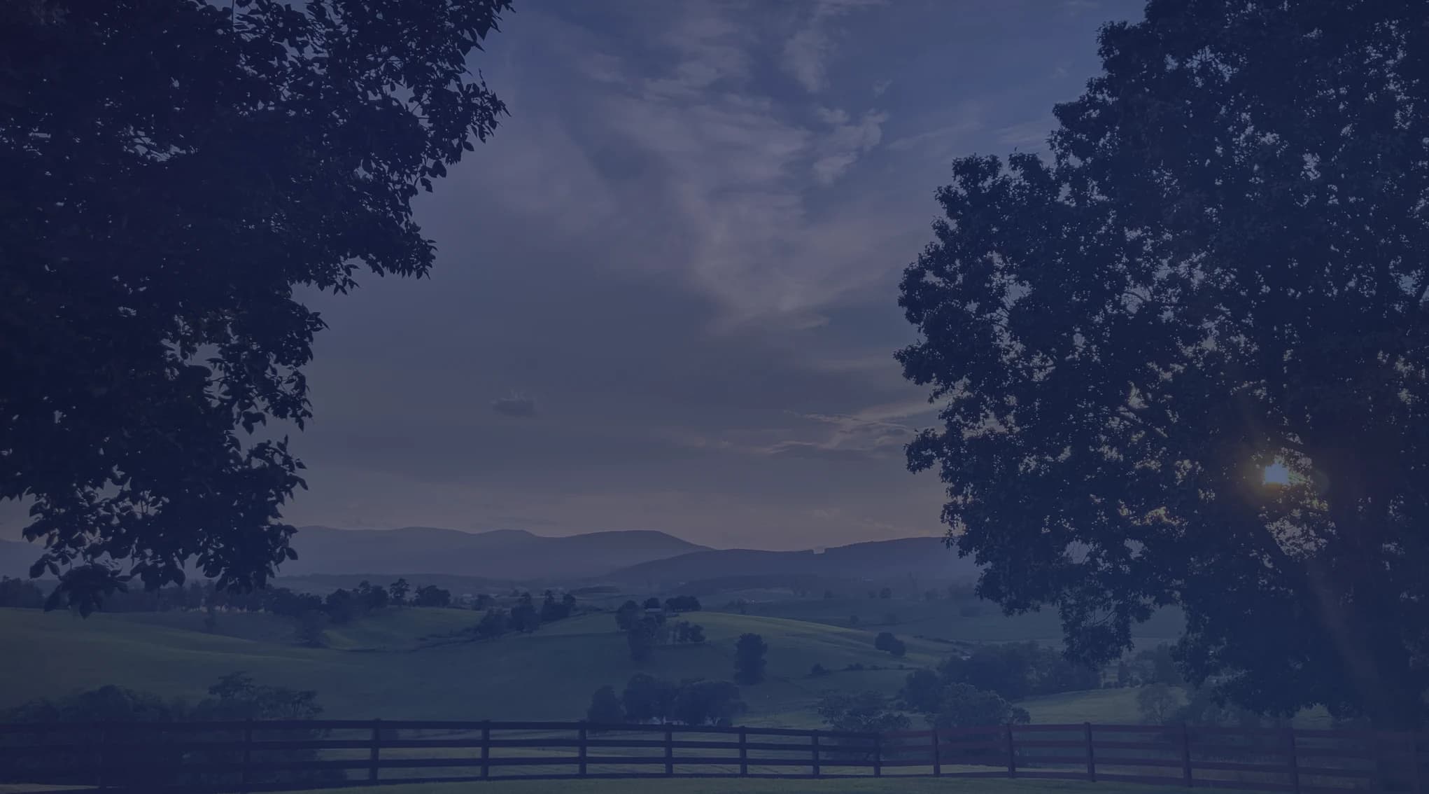 Rolling Shenandoah Valley farmland behind a wooden fence in Virginia, framed by trees at dusk.