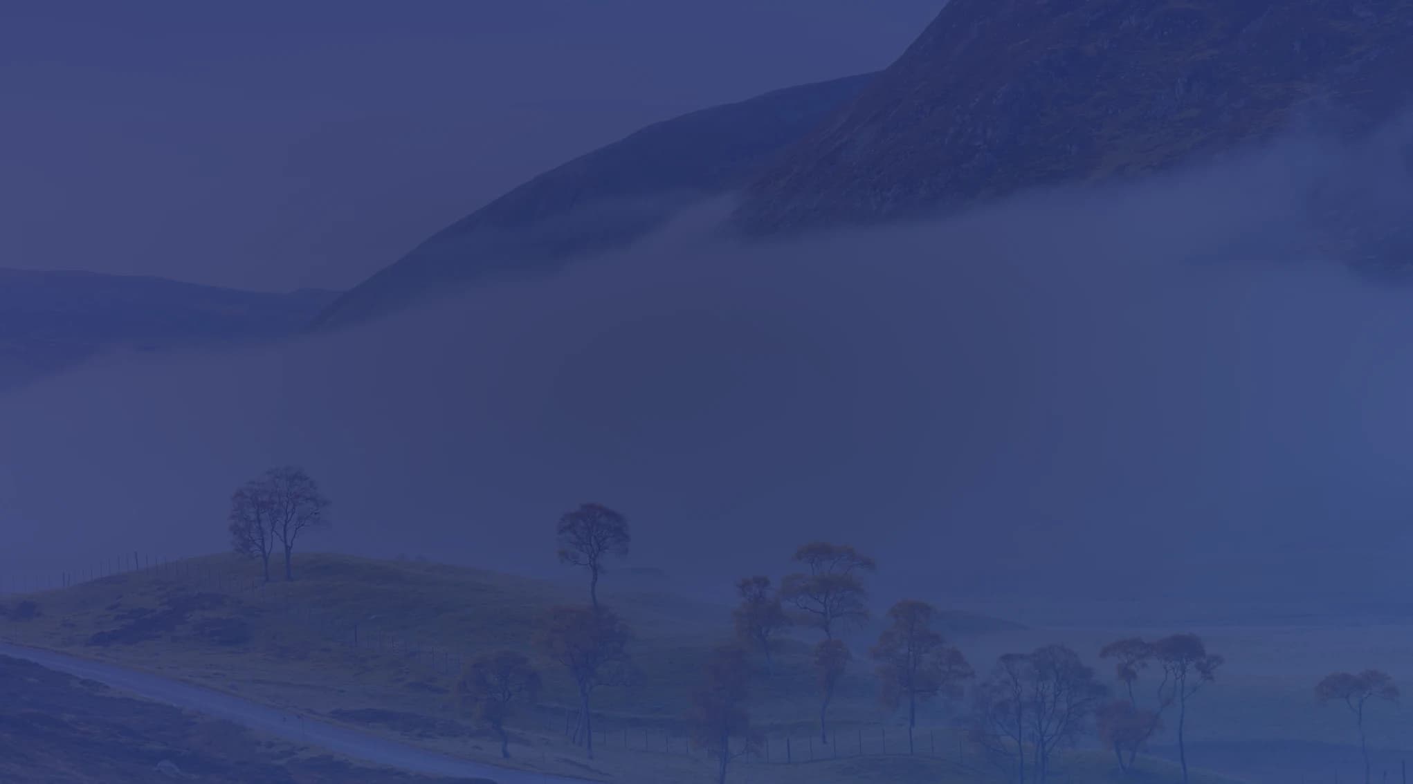 A low band of fog hangs across a bare mountainside above rolling hills dotted with autumn-colored trees and a winding road.