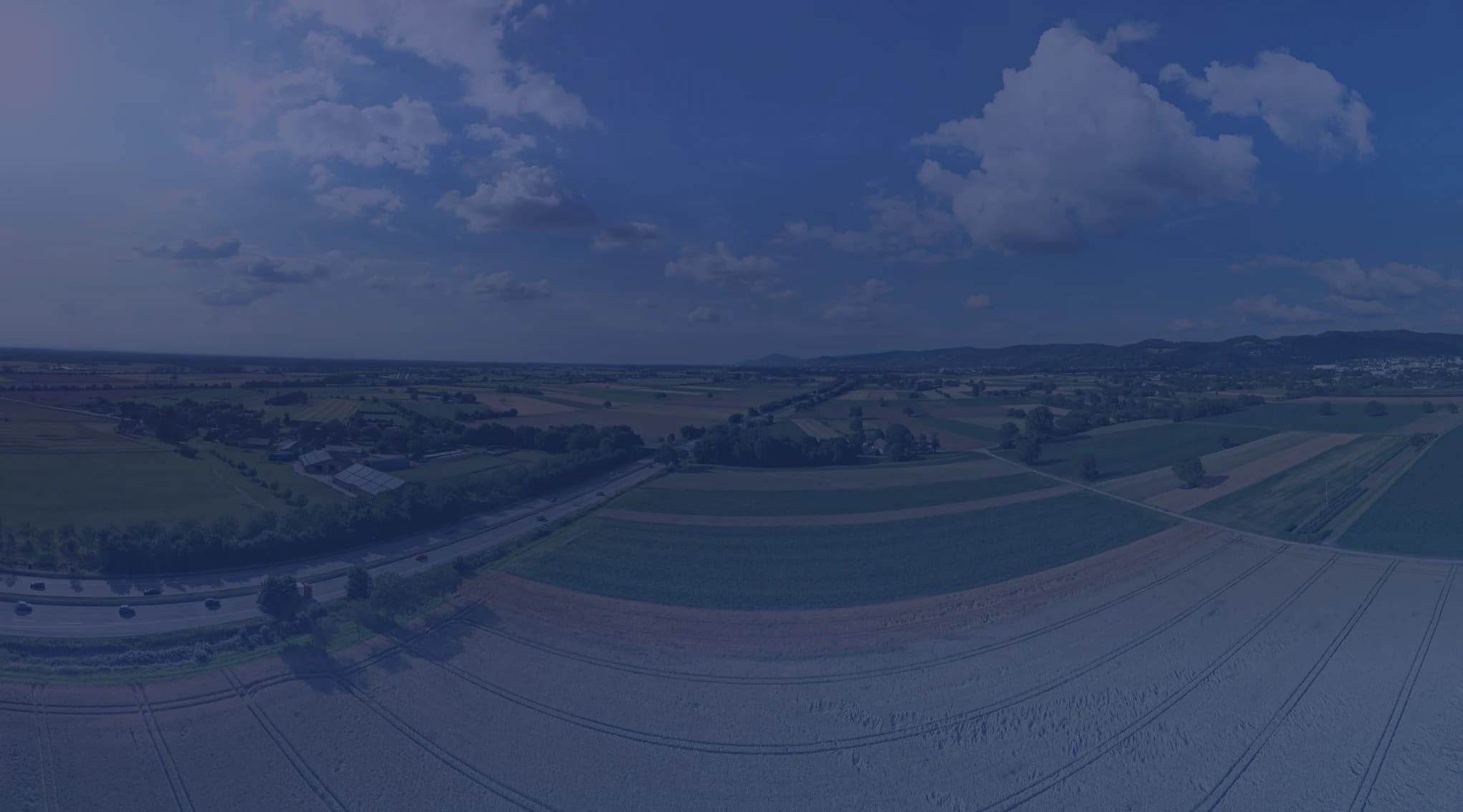 A wide aerial view of patchwork farm fields in green and gold, with low hills under a bright, partly cloudy sky.